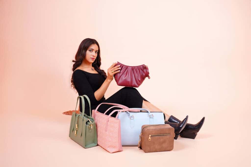 A woman in a black outfit sits on the floor holding a maroon handbag, surrounded by five other handbags in various colors: green, pink, light blue, brown, and light pink, against a pale background.