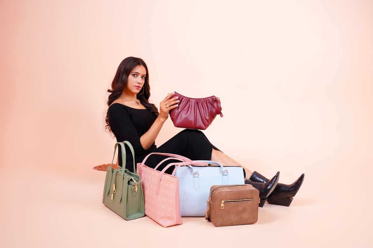 A woman in a black outfit sits on the floor holding a maroon handbag, surrounded by five other handbags in various colors: green, pink, light blue, brown, and light pink, against a pale background.