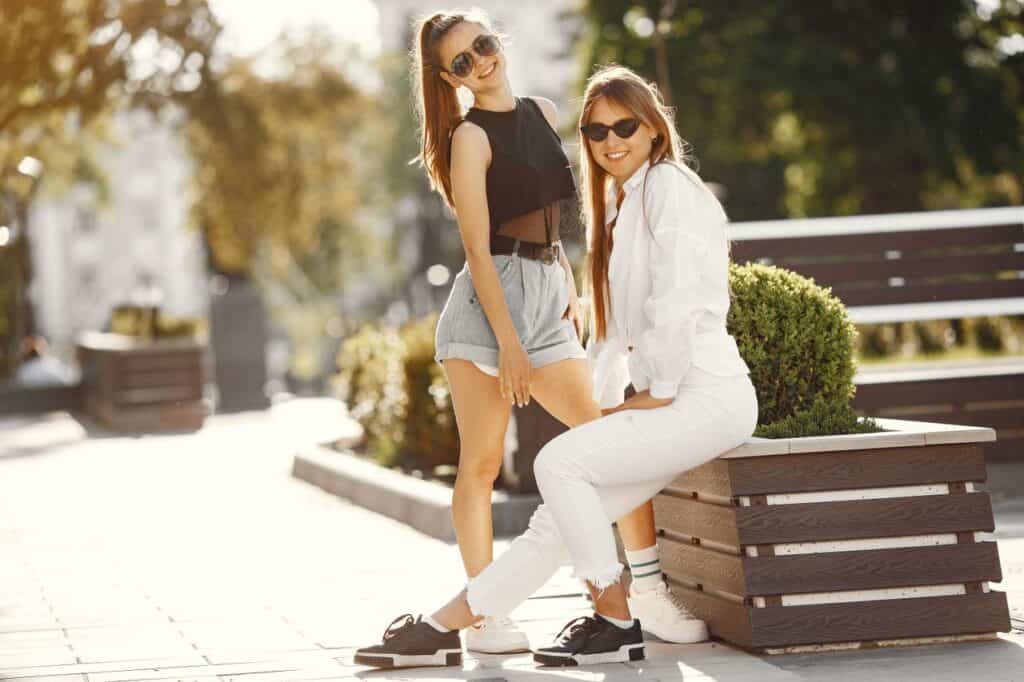 Two young women outdoors on a sunny day, one standing in shorts and a black top, the other sitting on a bench planter in white pants and a shirt. Both are wearing sunglasses and smiling.