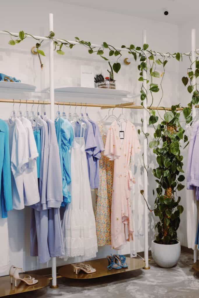 Clothing rack with pastel-colored shirts, dresses, and blouses in shades of blue, white, and pink. Shoes are on the bottom shelf, and leafy green plants decorate the rack and floor. The setting is bright and stylish.